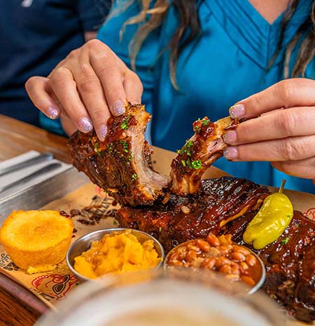 Woman pulling apart BBQ ribs on a tray with cornbread, sweet potato casserole, and baked beans at Lizzie's Memphis-Style BBQ